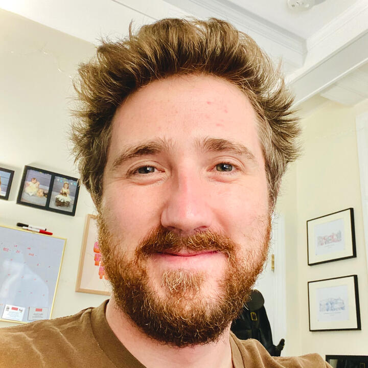 RJ Foley with a relaxed smile on his face. His amber beard and hair are a mix of slightly dirty blonde and golden auburn, styled naturally with a flip at the front of his scalp which complements his teal shirt.